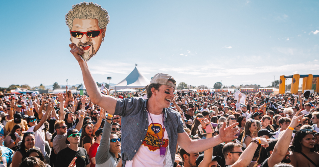 A person on shoulders in a concert crowd holding a Guy Fieri cutout
